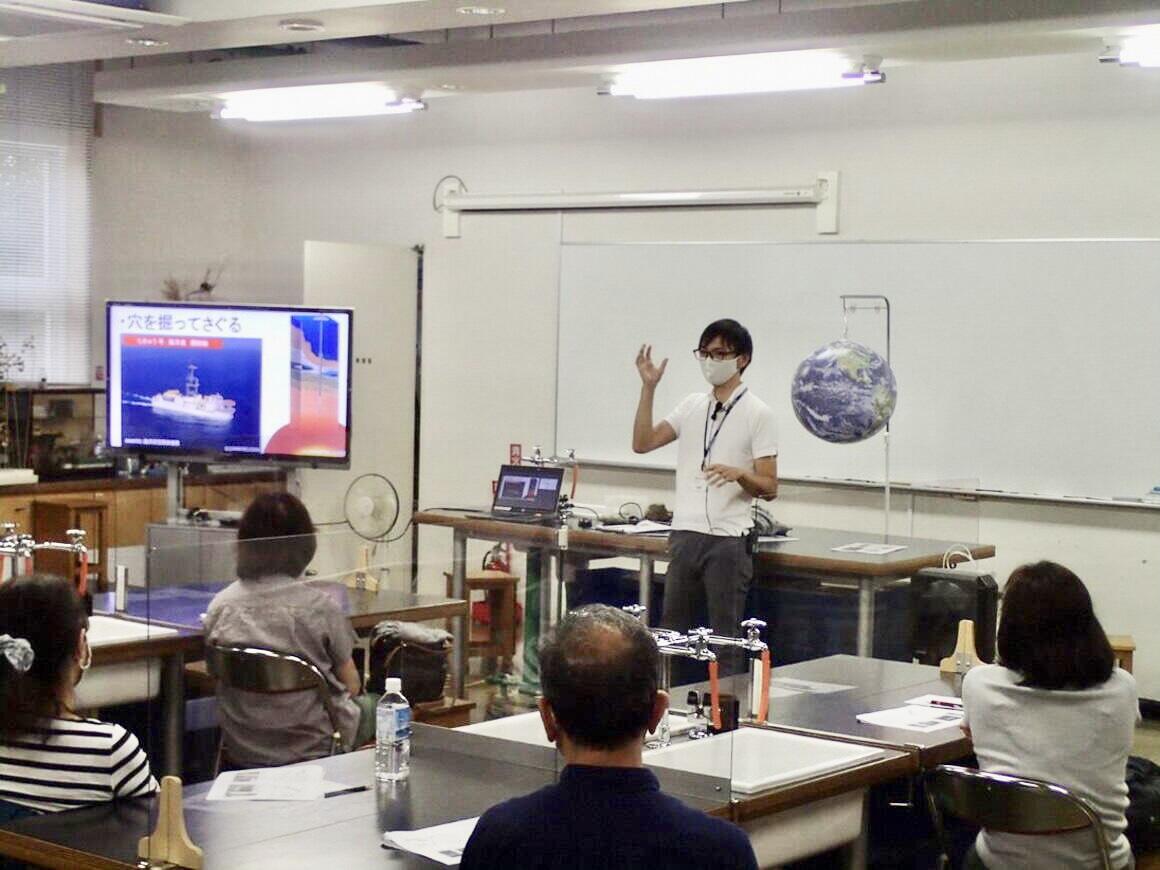 地球科学入門の講座の様子。左側にモニター、中央にマスク姿の講師、右側に地球儀が浮いています。