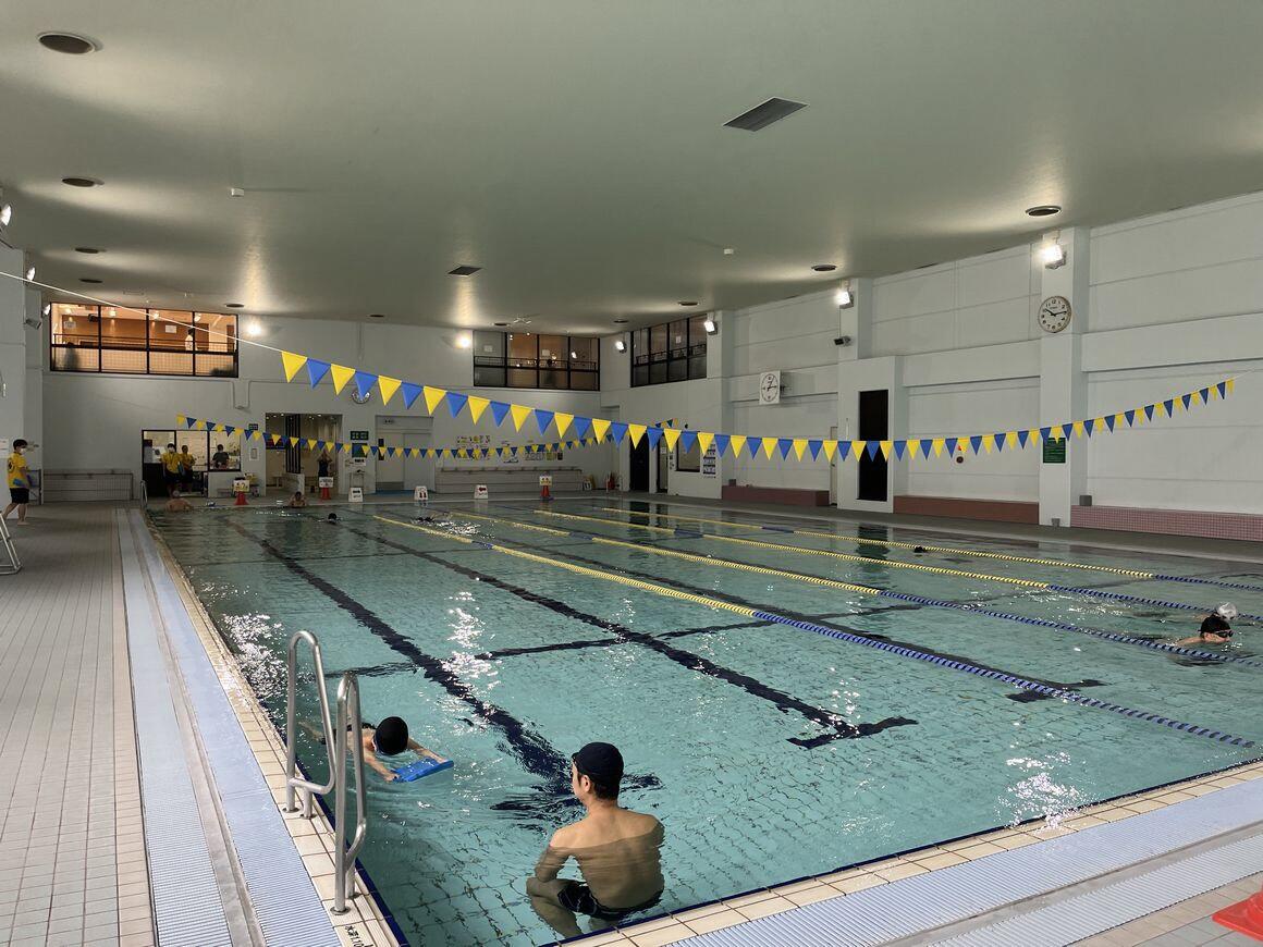 りゅうせんえんの温水プールの写真。25メートルの一般用プールで、ビート板を使って子供がバタ足の練習をして、プールの中で大人が見守っています。