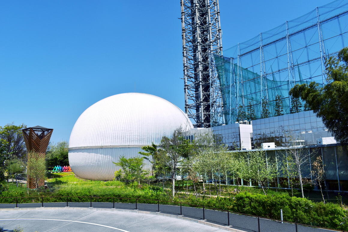 多摩六都科学館の外観の写真。中央に白く輝くサイエンスエッグ、右側に銀色の展示室があります。後ろにはスカイタワー西東京があります。