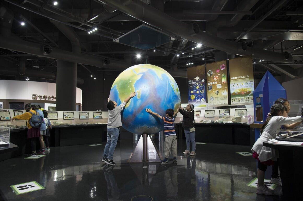 地球に関する展示の中央で、三人の子供が両手を広げて、大きな地球の模型に触っている写真