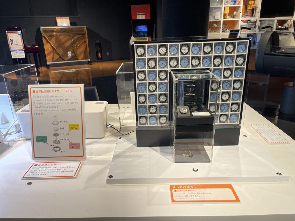 光発電時計のしくみについての展示の写真。右側には、小さな青い時計と白い時計が交互に七段に並べられています。